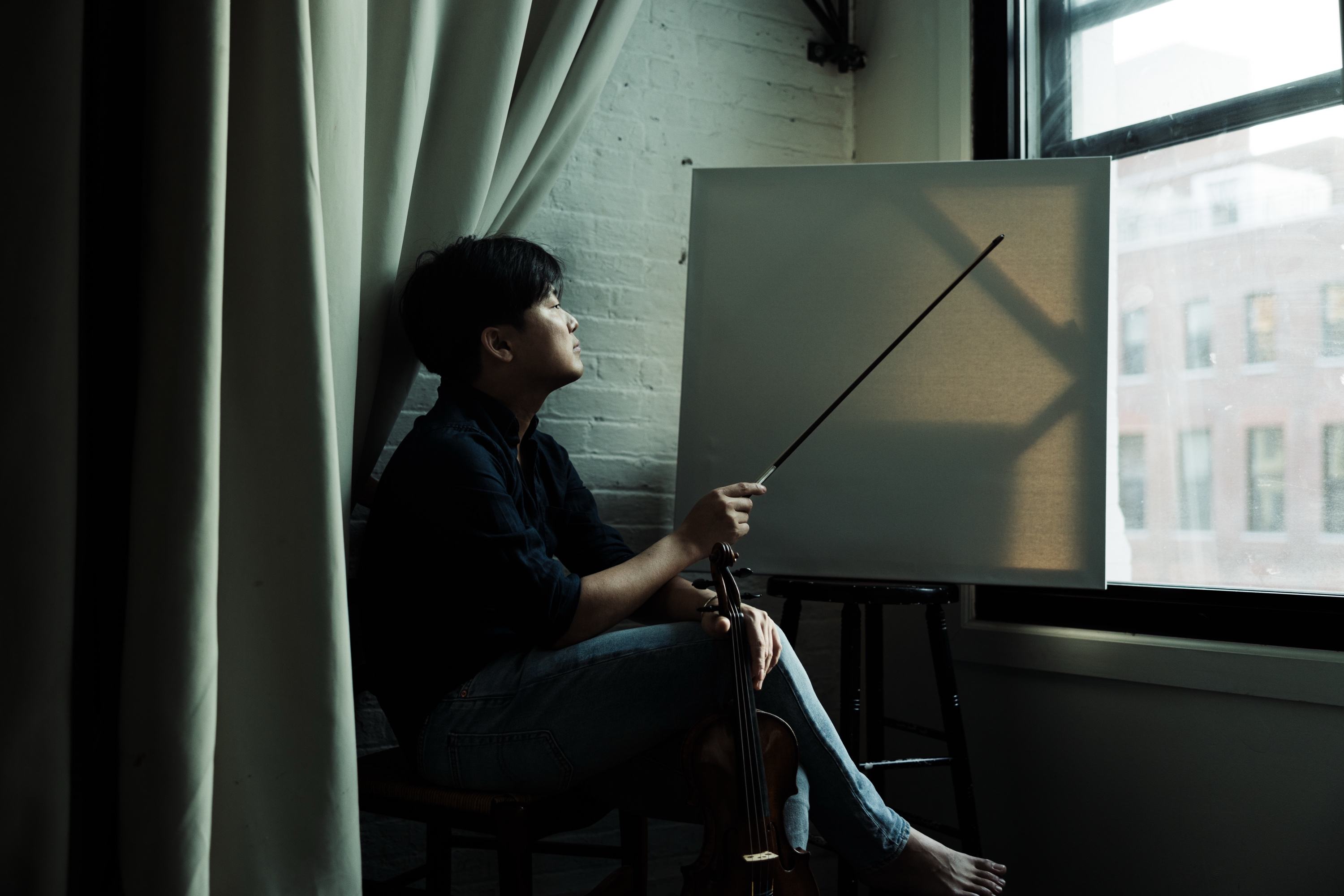 Siwoo Kim seated by a loft window, raising his bow toward a blank canvas